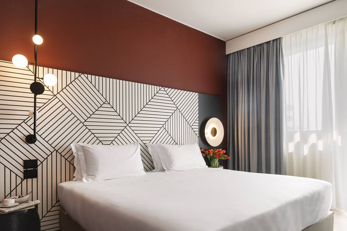 Moderni hotellihuone, jossa on geometrisesti kuvioitu sängynpääty, siististi pedattu sänky valkoisilla lakanoiden kanssa, yöpöytä lampulla ja ikkuna, jossa on läpikuultavat verhot.