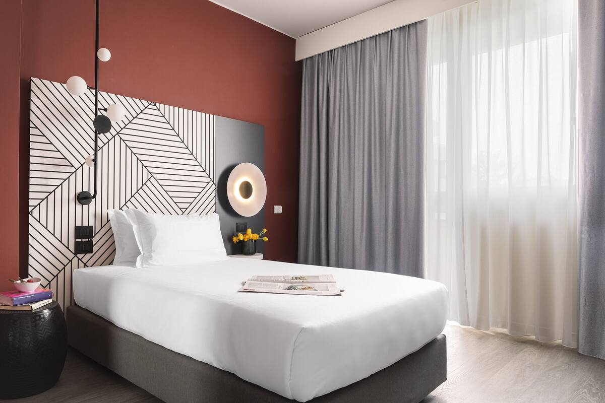 Moderni hotellihuone, jossa on suuri sänky, geometrisesti kuvioitu pääty, yöpöytä lampulla ja näkymä ulos ohuiden verhojen läpi.