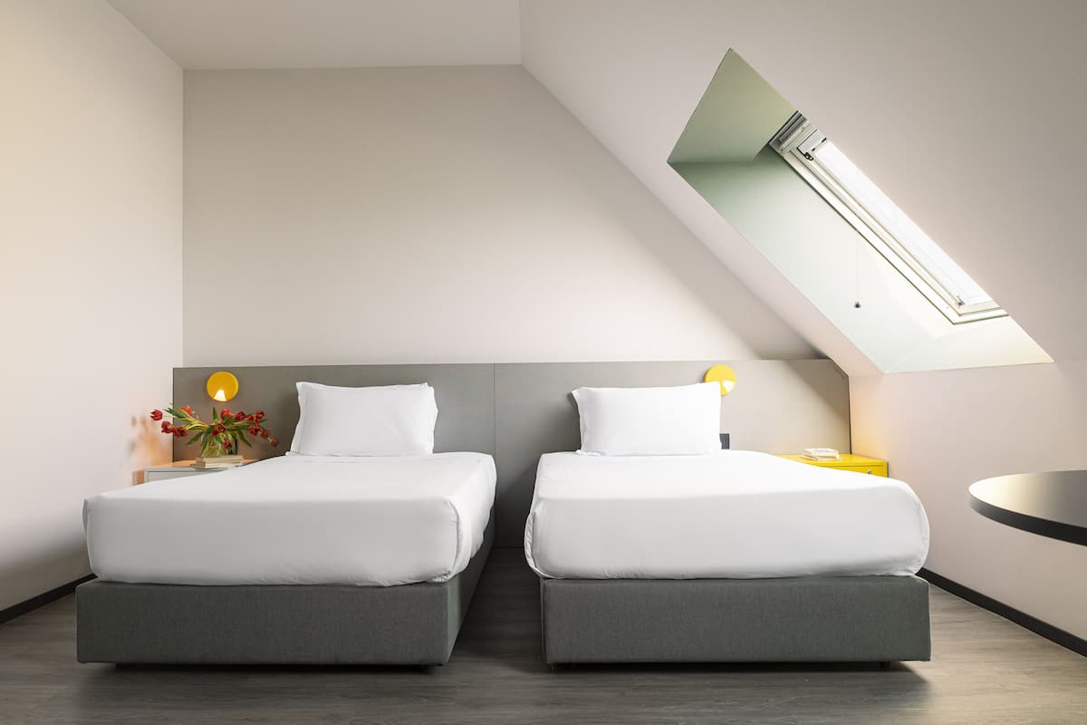 Moderni hotellihuone, jossa on kaksi sänkyä, kattovalokuvio ja pieni pyöreä pöytä.