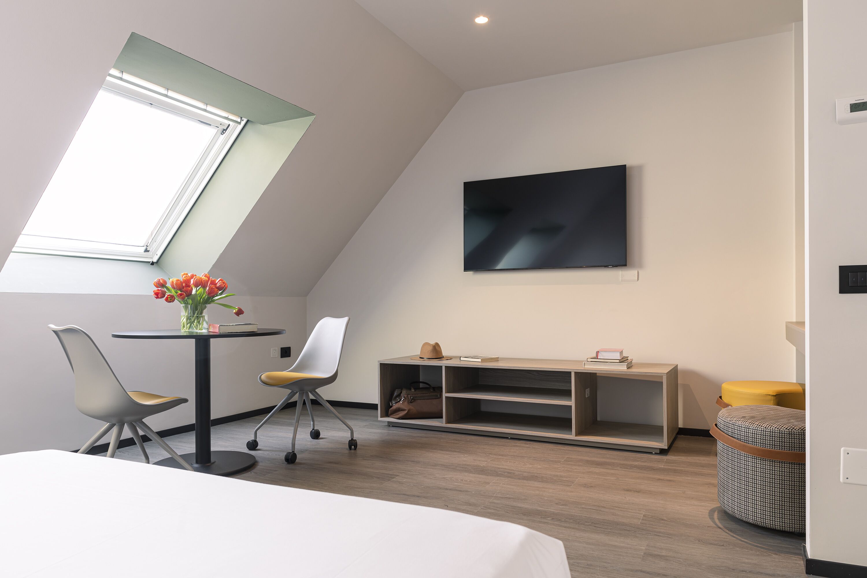 Et moderne hotelværelse med skråvæg, stort vindue, fladskærms-tv, rundt spisebord, to stole, et lille sofabord og en sofa.