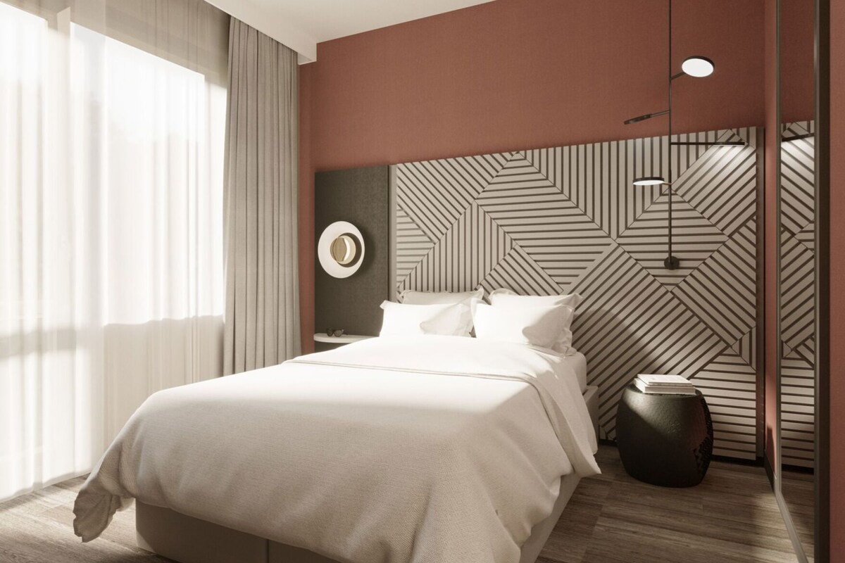 A modern hotel room with a large bed, a round mirror, a geometric-patterned headboard, and a large window with curtains.
