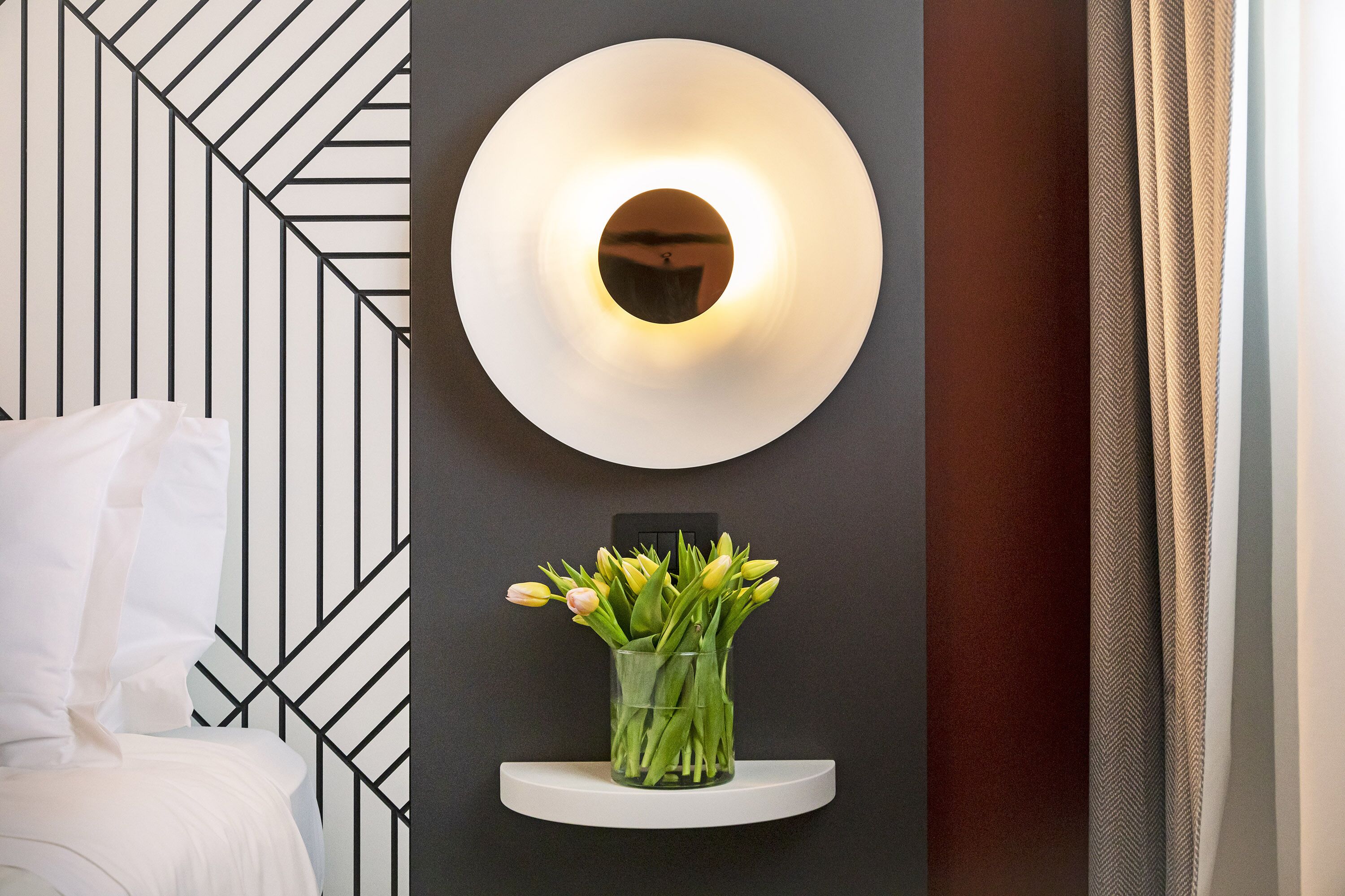 Una habitación moderna con una pared de diseño geométrico, una lámpara redonda de pared y un florero de vidrio con tulipanes amarillos sobre un estante blanco.