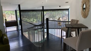 Een modern interieur met een trap naar een balkon, een eethoek met een tafel en stoelen, en grote ramen met uitzicht op groen.