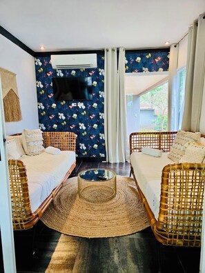 Een kamer met twee rieten banken, een rond geweven salontafel, een flatscreen televisie aan een wand met bloemenprint, en een raam met witte gordijnen.