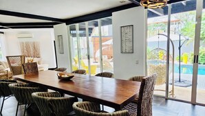 Een moderne eetruimte met een houten tafel en geweven stoelen, grote ramen en uitzicht op een zwembad en terras.