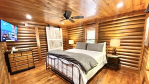 A wooden cabin bedroom with a bed, bedside tables, a ceiling fan, and a TV on the wall.