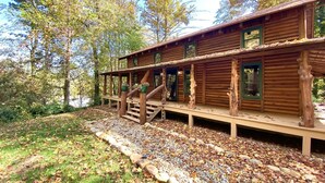 A wooden cabin with a deck, surrounded by trees and a gravel path.