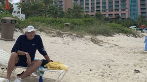 A beach scene with a man sitting on a lounge chair, a palm tree, and a large hotel building in the background. - Luxurious Atlantis Harborside resort beauty 3 bedroom deluxe Villa (Nassau)