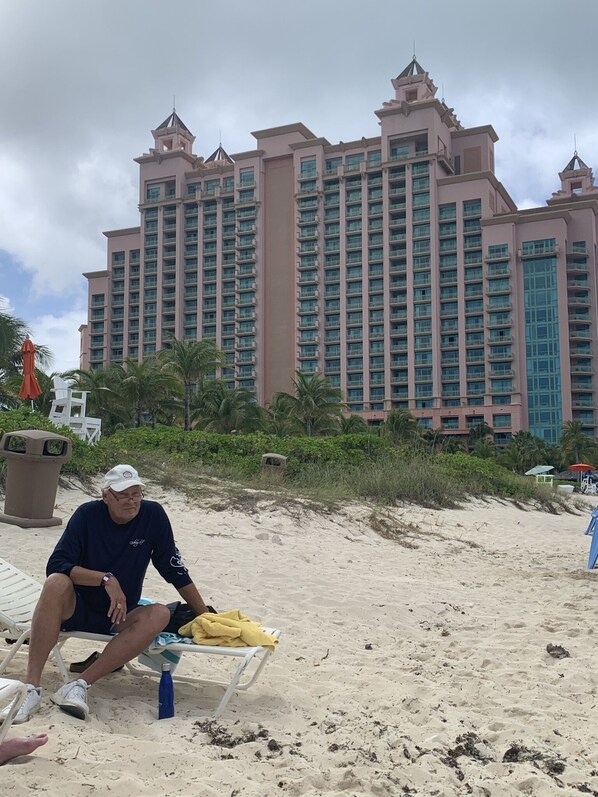 A beach scene with a man sitting on a lounge chair, a palm tree, and a large hotel building in the background.