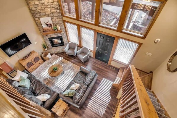 A spacious living area with a stone fireplace, a large sofa, and a television. There are multiple windows providing natural light and a view of the outside.