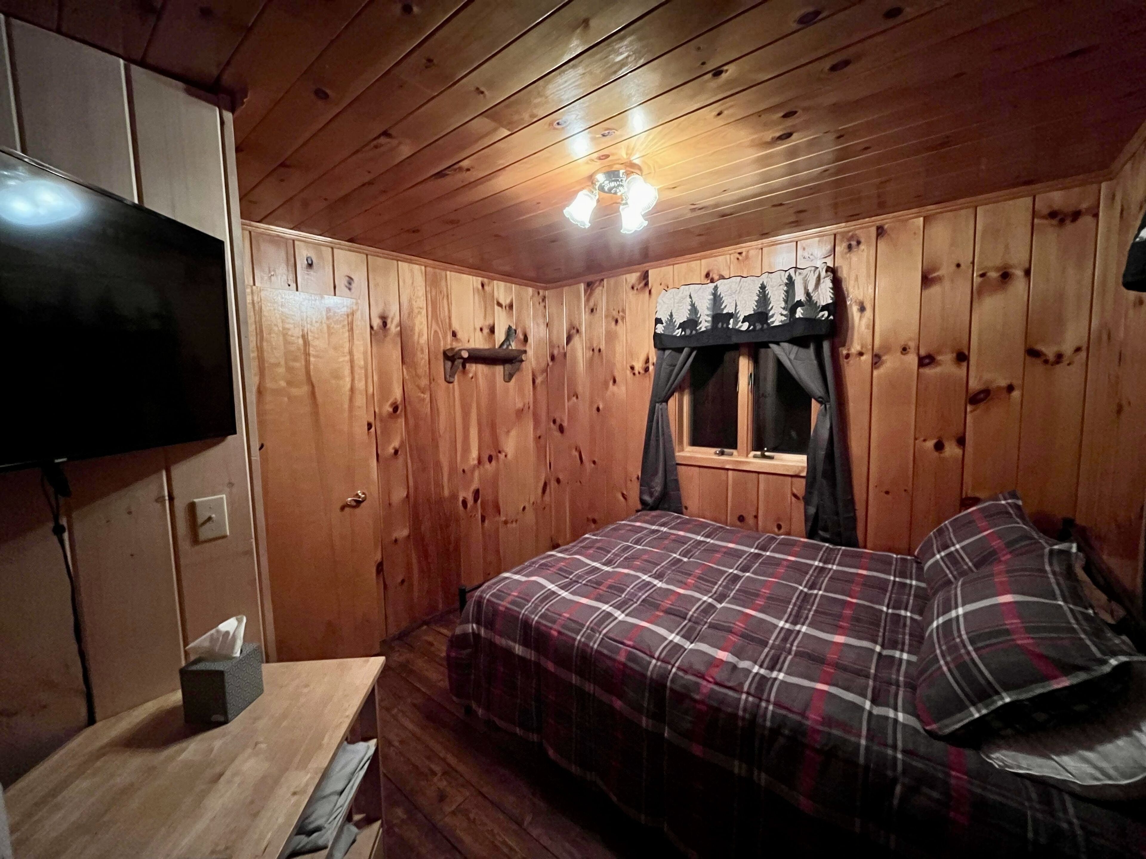 A wooden cabin room with a bed, a mounted TV, and a window with curtains.