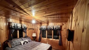 A cozy wooden cabin bedroom with a bed, curtains, a lamp, and a window. - Cozy Errol Home conveniently close to town and with snowmobile access!  (Errol)