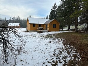 A wooden cabin with a porch, surrounded by snow-covered ground and trees.