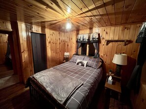 A wooden cabin bedroom with a bed, bedside lamps, and a window with curtains.