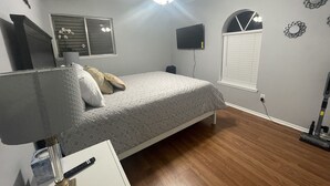 Un dormitorio con cama, ventilador de techo, televisor, ventana con persianas y aspiradora.