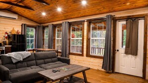 Interior acogedor de una cabaña con un sofá, una mesa de madera y ventanas grandes con cortinas.