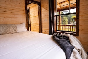 Habitación de cabaña de madera con una cama, una ventana que da a una terraza de madera y una puerta que sale al exterior.