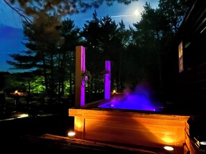A wooden hot tub with a purple light, surrounded by trees and a clear sky.