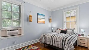 Une chambre à coucher comprenant un lit, deux tables de chevet, une fenêtre avec stores, un climatiseur fixé au mur et un tapis coloré.