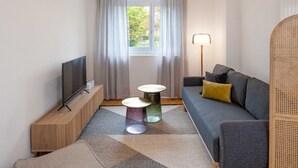 Een moderne woonkamer met een flatscreen televisie op een houten standaard, een grijze bank met een geel kussen, twee kleine bijzettafeltjes en een vloerlamp.