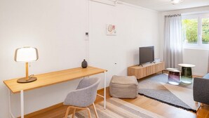 Een moderne, goed verlichte kamer met een houten bureau, een grijze stoel, een televisie op een houten standaard, een klein rond tafeltje, en een raam met gordijnen.