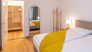 Een hotelkamer met een bed, nachtkastje met een lamp, spiegel, kledingkast en een houten vloer.