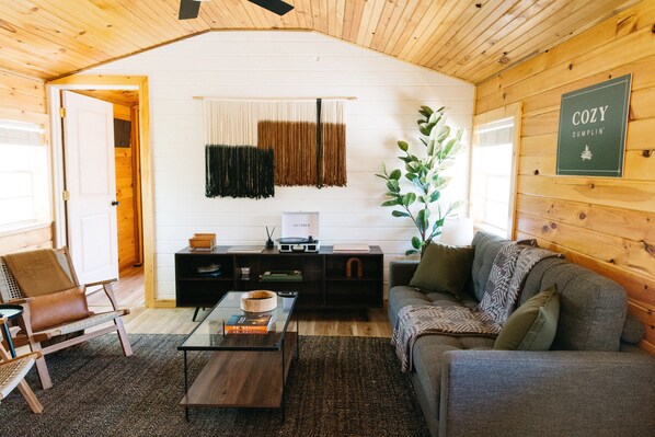 A cozy living room with a wooden ceiling, a sofa, a coffee table, and a bookshelf. - Cozy Dumplin' RRG - Warm & Modern Stay in RRG/Auxier (Pine Ridge)