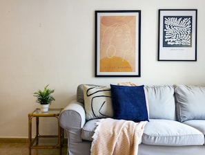 Uma sala de estar com um sofá cinzento, uma mesinha de apoio com uma planta e duas obras de arte emolduradas na parede.