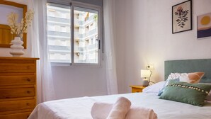 A hotel room with a bed, a wooden dresser, a nightstand with a lamp, and a window with blinds. - Pink Sunshine beach apartment (Torrevieja)