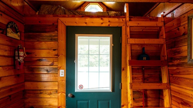 Interior de una cabaña con puerta verde, paredes de madera, una ventana, un banco de madera y un bote de basura.