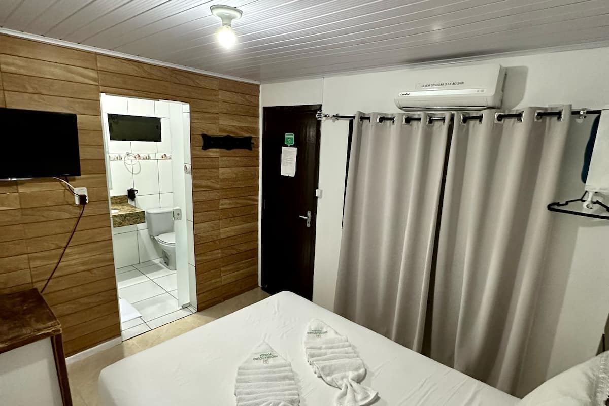 Quarto de hotel com cama, televisão, banheiro visível por uma porta aberta e um aparelho de ar condicionado na parede.