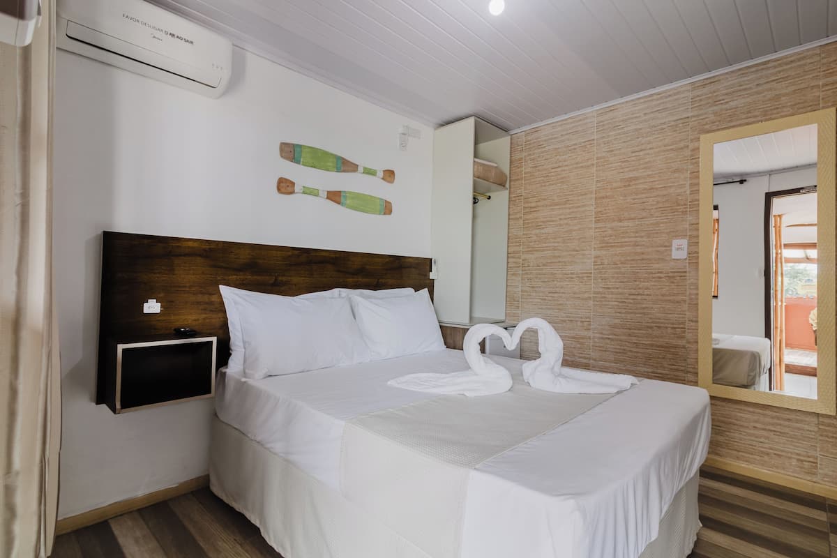 Cama bem arrumada com lençóis brancos, cabeceira de madeira e ar condicionado fixado na parede.