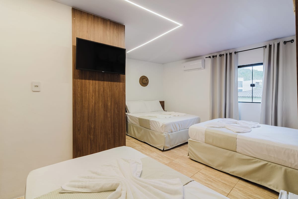 Quarto de hotel com duas camas, uma televisão de tela plana na parede, uma janela com cortinas e um painel de madeira na parede à esquerda.