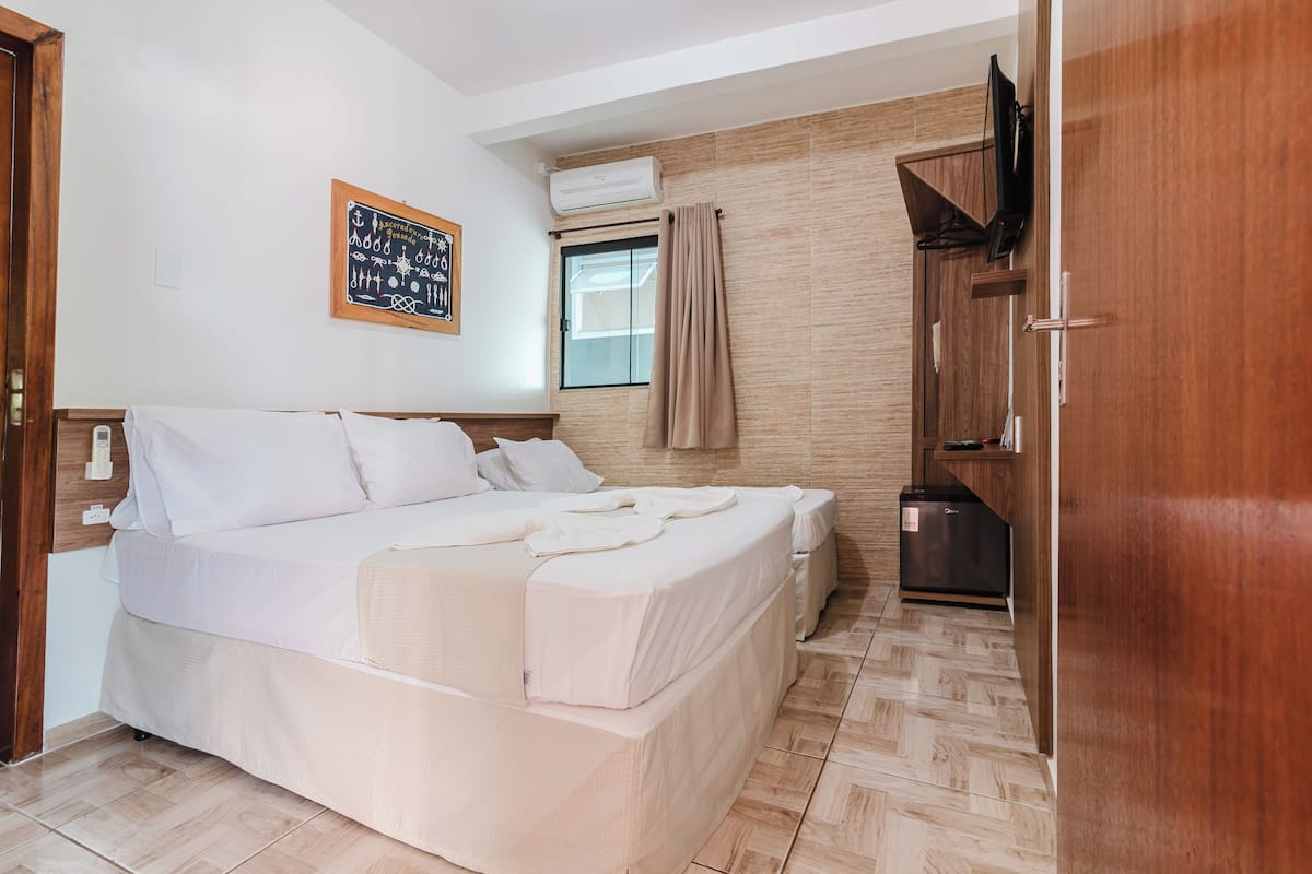 Quarto com cama, televisão, janela com cortinas e um pequeno espaço com cozinha.