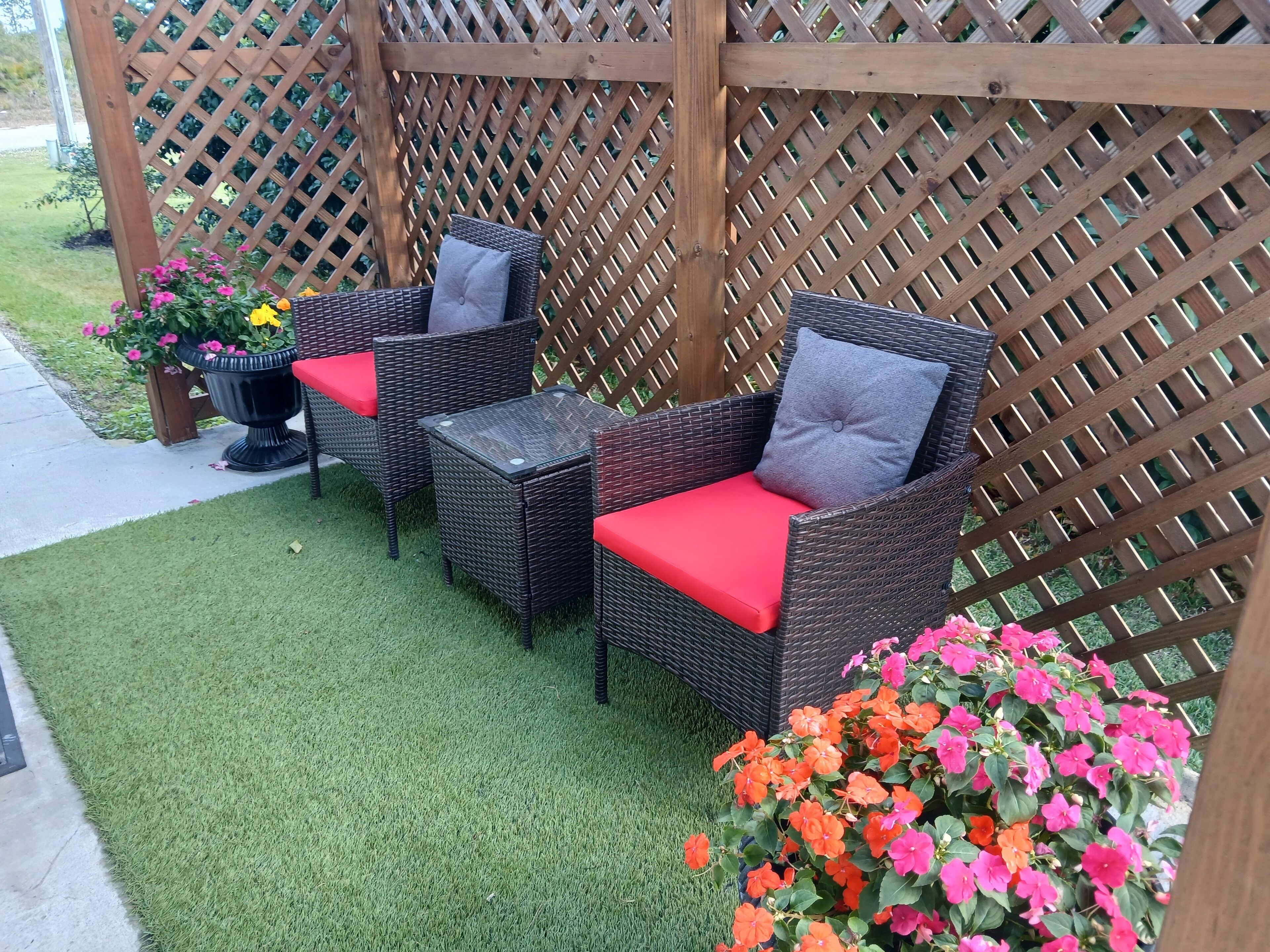 An outdoor seating area with wicker chairs and a glass table, surrounded by a wooden lattice fence and potted plants.