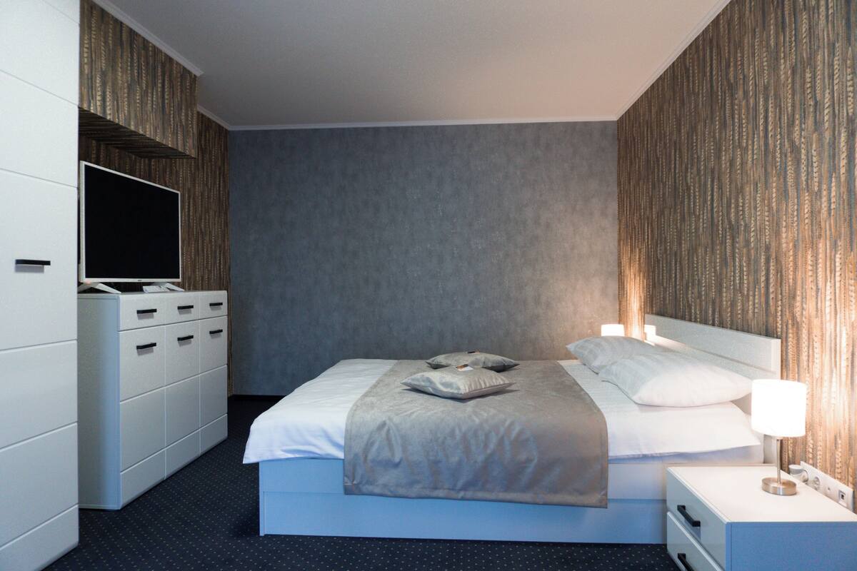 A modern hotel room with a large bed, a flat-screen TV, a dresser, and a bedside table with a lamp.