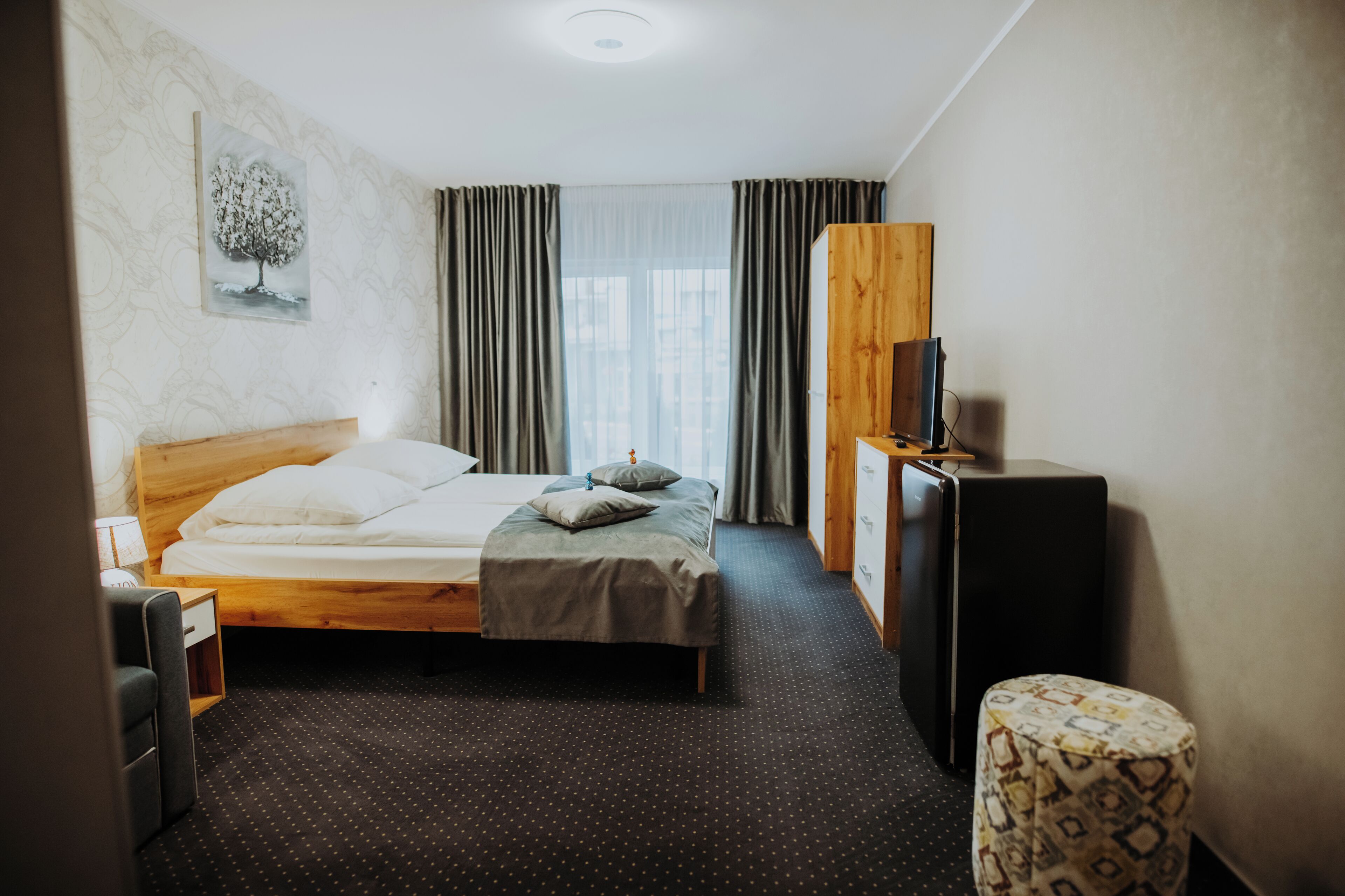 A hotel room with a wooden bed, a small bedside table, a TV, and a patterned ottoman.