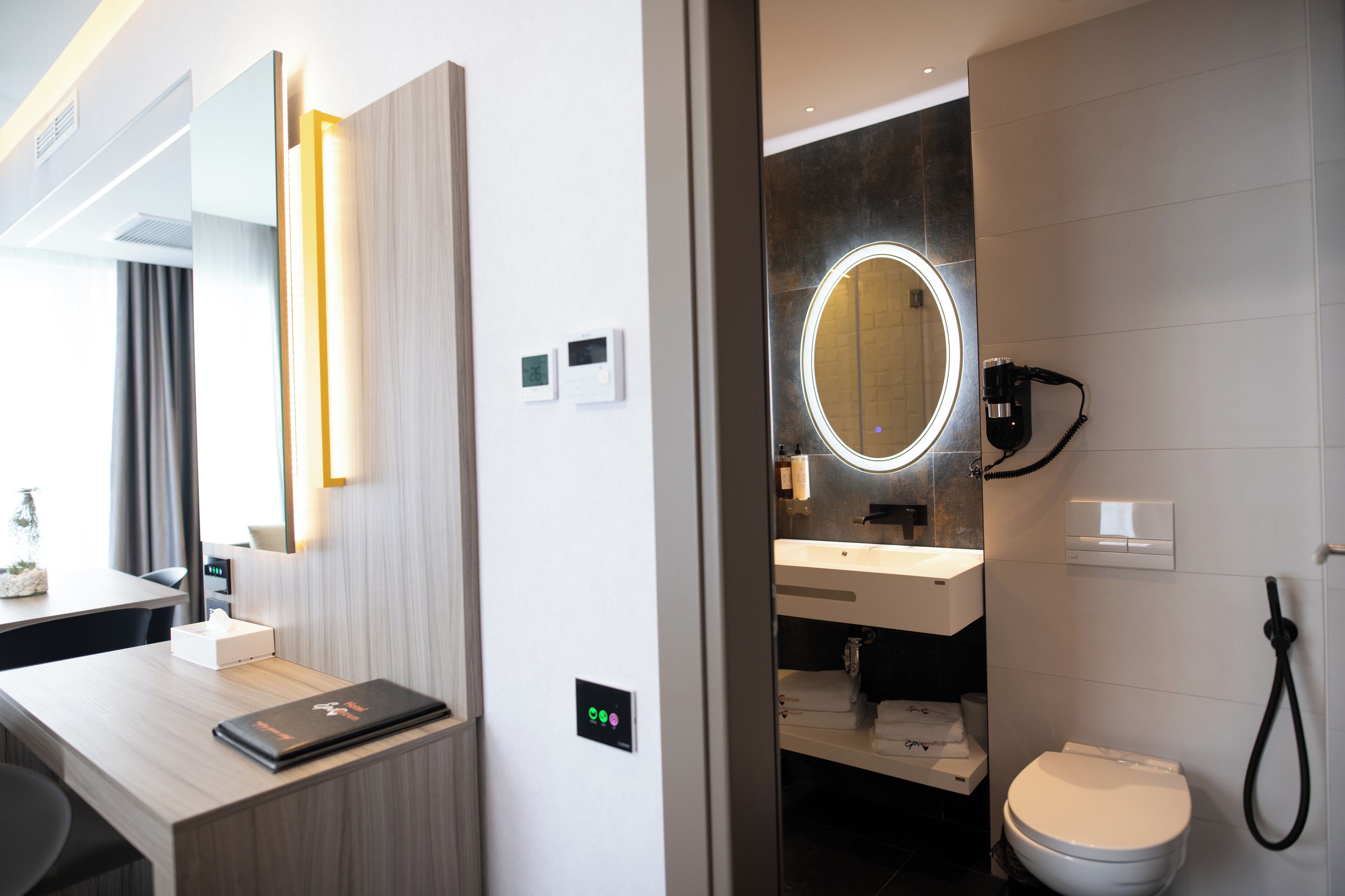 A modern hotel room with a wooden desk, a laptop, a mirror with a light, a toilet, and a shower.