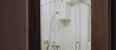 A glass-enclosed shower with a white marble wall, a white toilet, and a small shelf above the toilet.