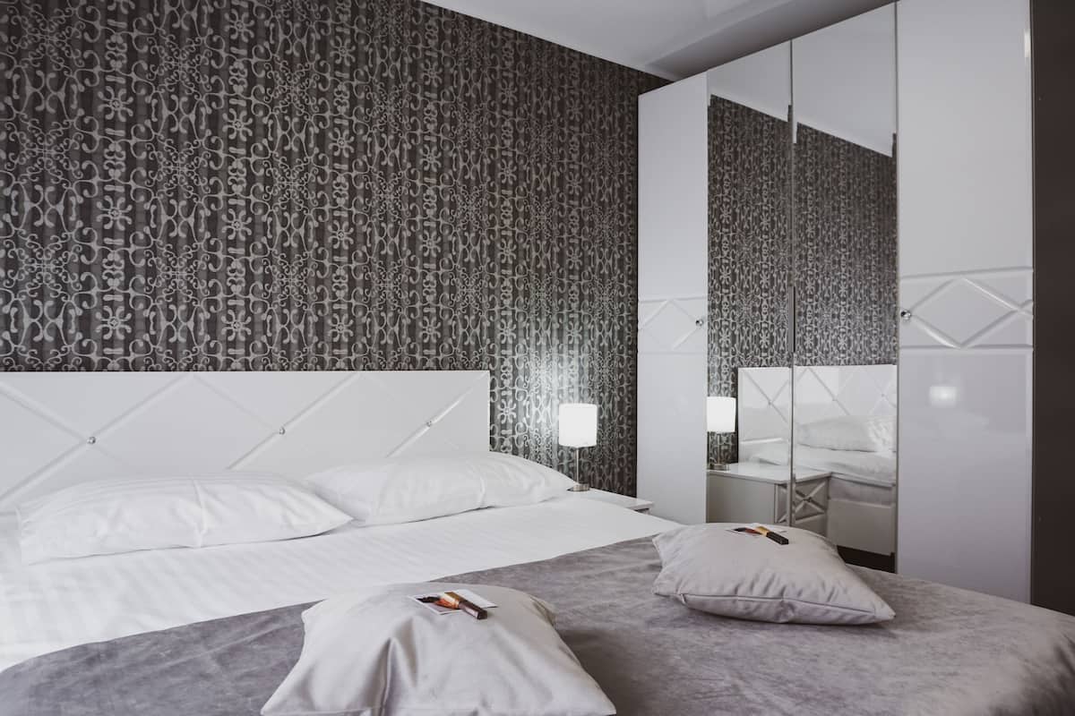 A modern bedroom with a patterned wallpaper, a tufted headboard, a large mirror, and a bedside table with a lamp.