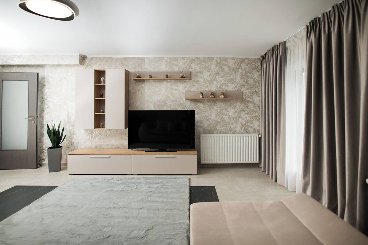 A modern living room with a flat-screen TV, built-in shelves, a potted plant, and a large window with curtains.