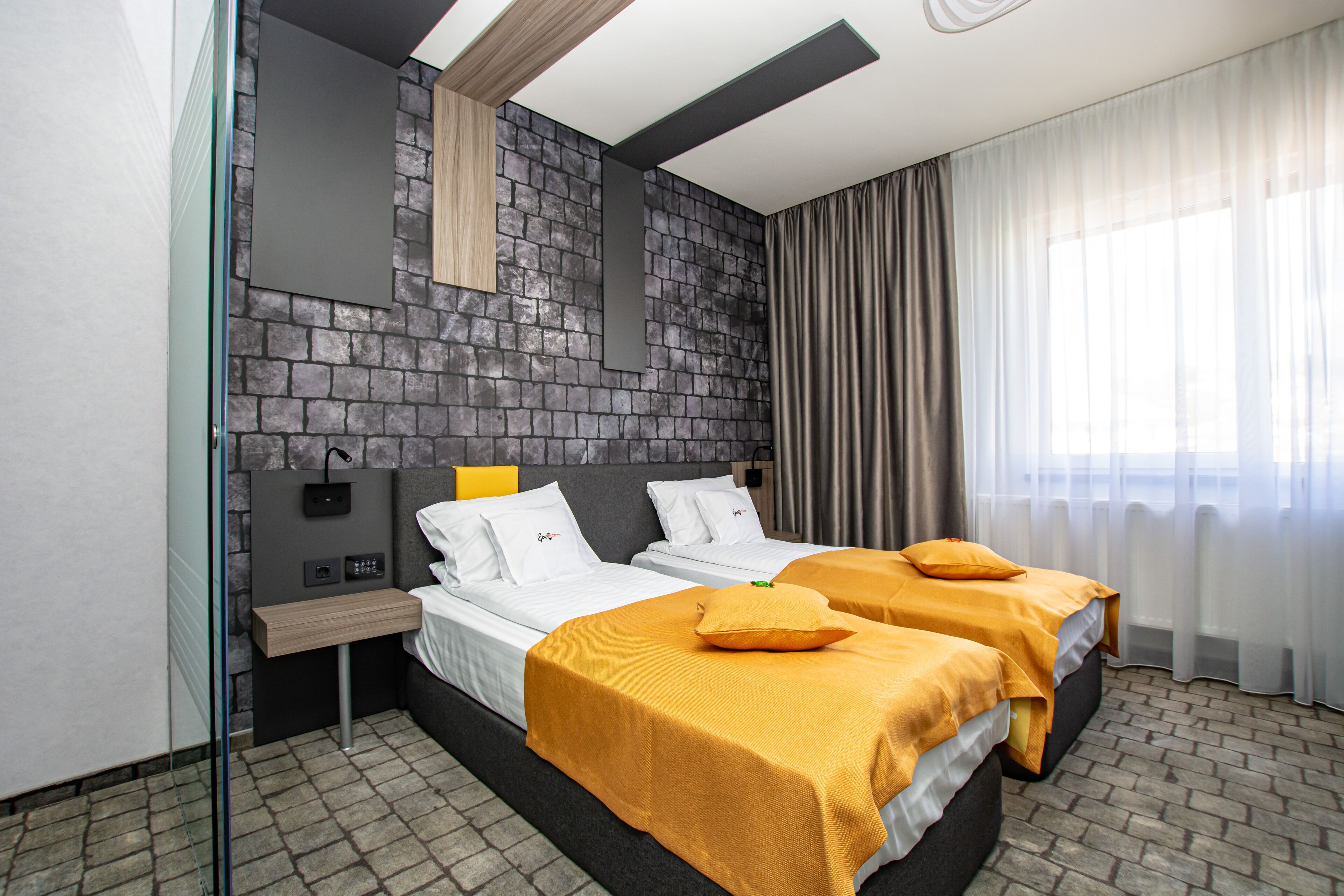 A modern hotel room with two beds, a stone accent wall, a bedside table, and a large window with curtains.