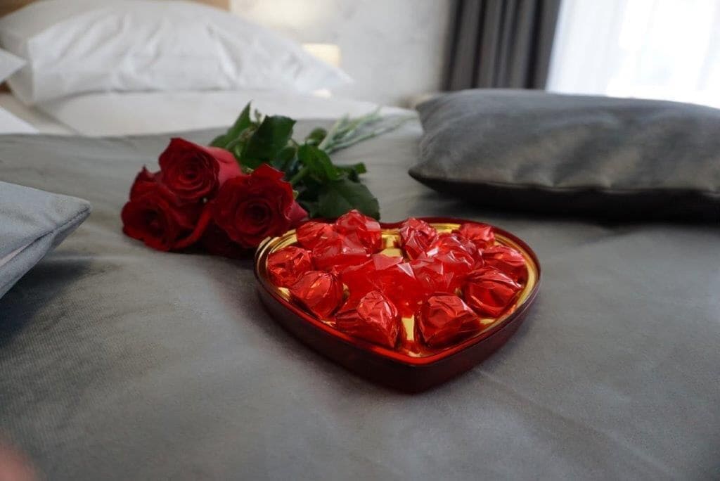 Una cama con una caja de bombones con forma de corazón y dos rosas rojas.