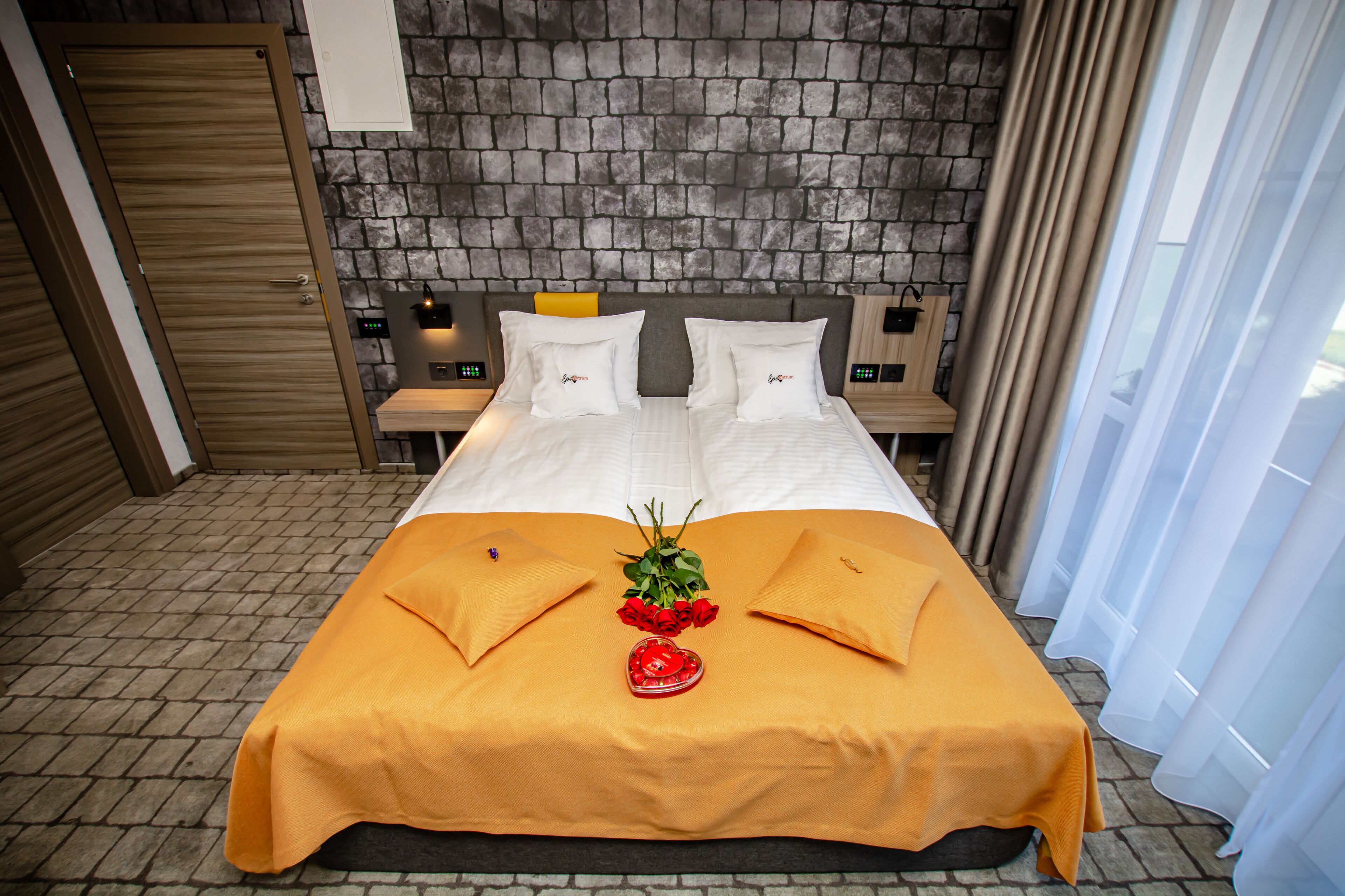 Habitación de hotel con pared de ladrillo, cama con sábanas blancas, dos almohadas, mesita de noche con lámpara y ventana con cortinas translúcidas.