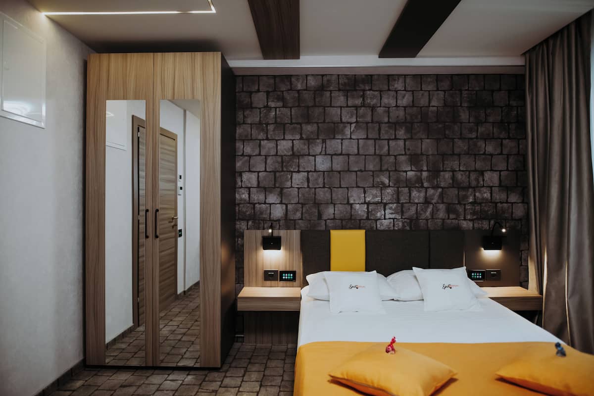 A hotel room with a brick-patterned headboard, a wooden wardrobe, a bedside table with a lamp, and a bed with white bedding and a yellow accent.