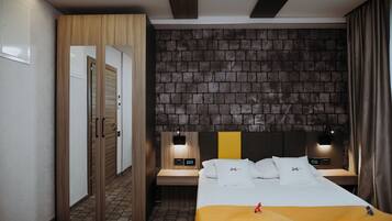 A hotel room with a brick-patterned headboard, a wooden wardrobe, a bedside table with a lamp, and a bed with white bedding and a yellow accent.