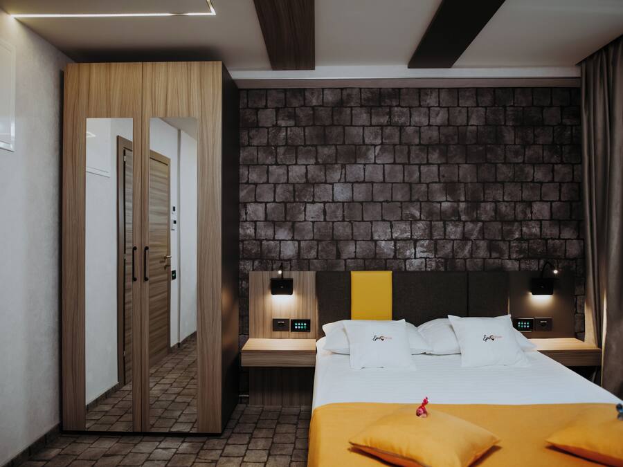 A hotel room with a brick-patterned headboard, a wooden wardrobe, a bedside table with a lamp, and a bed with white bedding and a yellow accent.