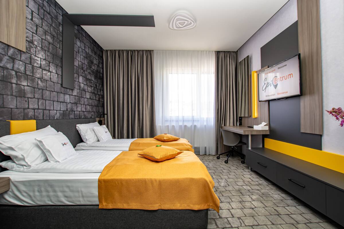 A hotel room with two beds, a desk, a TV, and a large window with curtains.