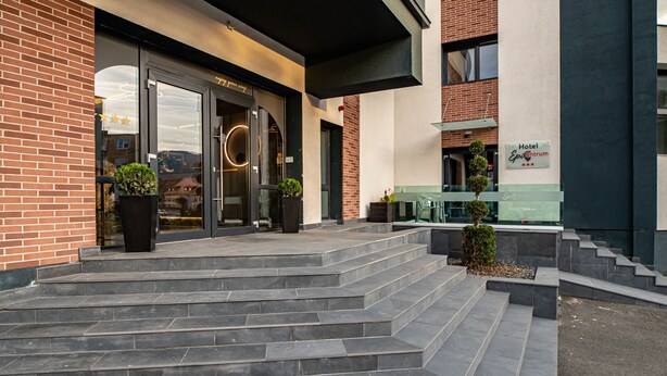 Entrada de un hotel con puerta de cristal, escalones que llevan a ella y un letrero que indica que es el Hotel Epsilon Atrium.