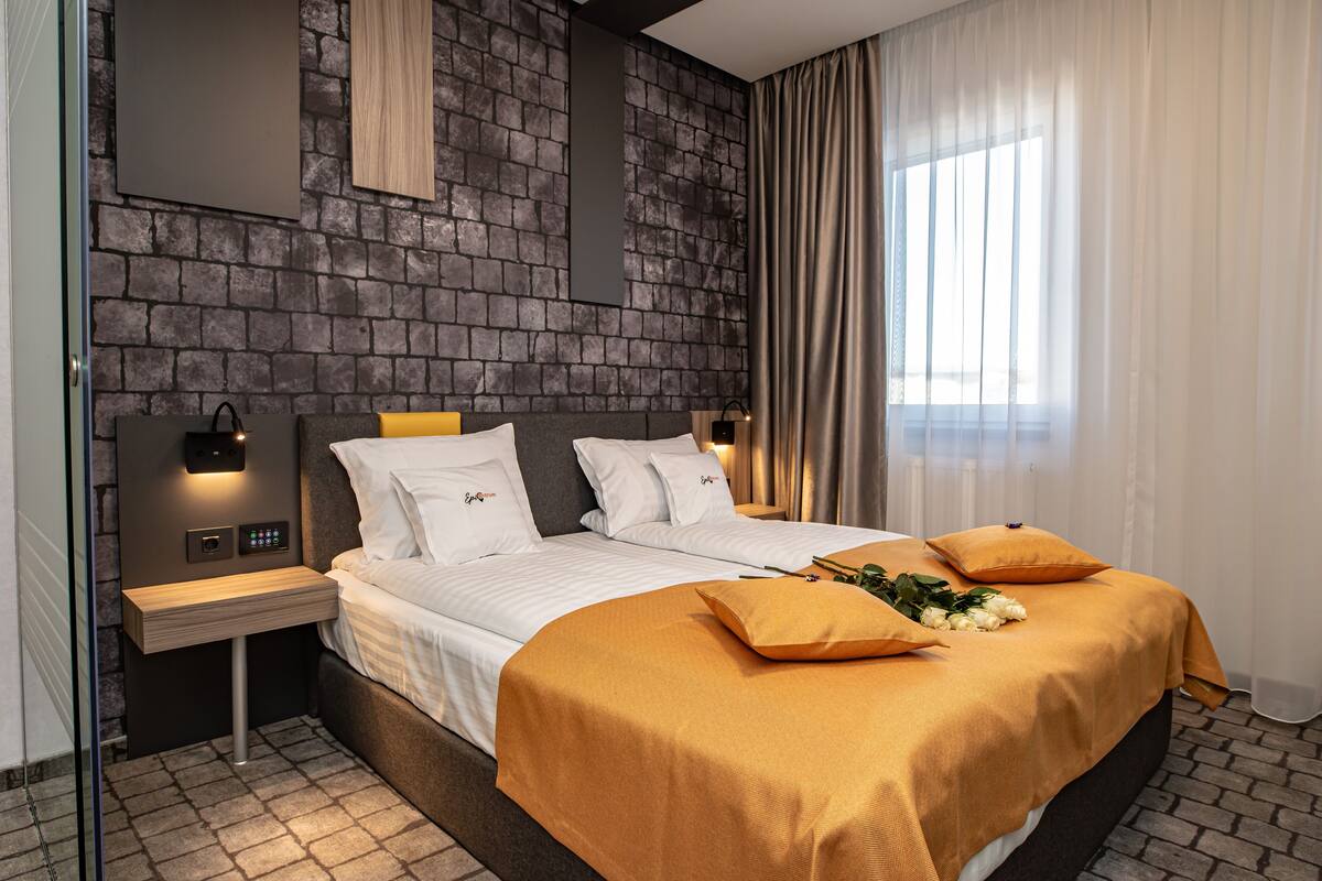 A modern bedroom with a stone wall, a large bed with white bedding and two orange pillows, a bedside table with a lamp, and a window with sheer curtains.
